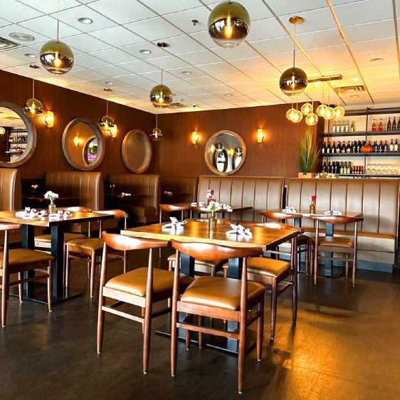 The image depicts a contemporary dining area with mid-century modern influences, featuring warm-toned wood framed chairs with cream cushions, complementing the wood-paneled walls and barrel-shaped mirrors. Overhead, chic spherical pendant lights add a golden glow to the inviting space.