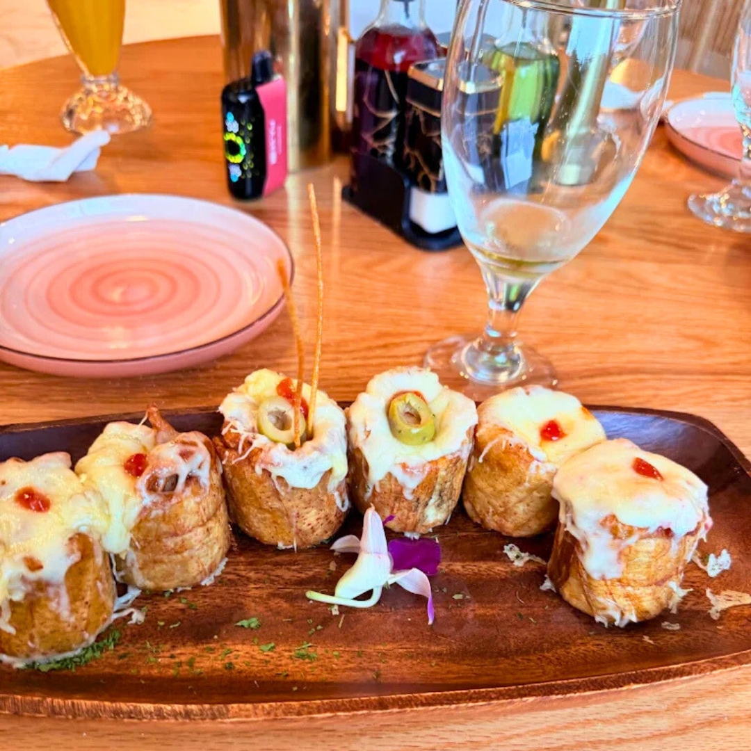 Cheese-topped stuffed potato rolls with olives on wood platter, resting on red-oak tabletop—Latin-inspired starter at La Juliana Miami.