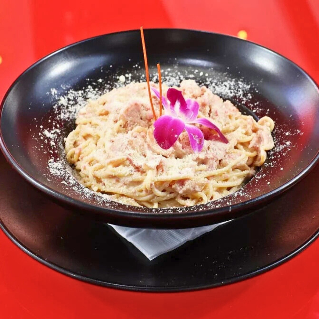 Creamy spaghetti carbonara topped with orchid, plated in black bowl on red-oak restaurant table at La Juliana Miami.