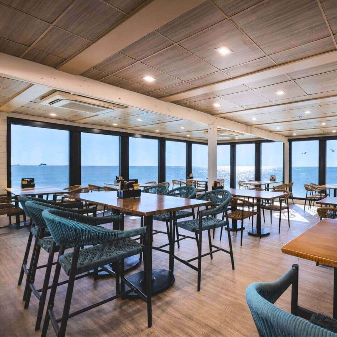 Waterfront dining space at Pier 62 Cocoa Beach showcasing turquoise rope chairs, hardwood floors, and expansive ocean-view windows